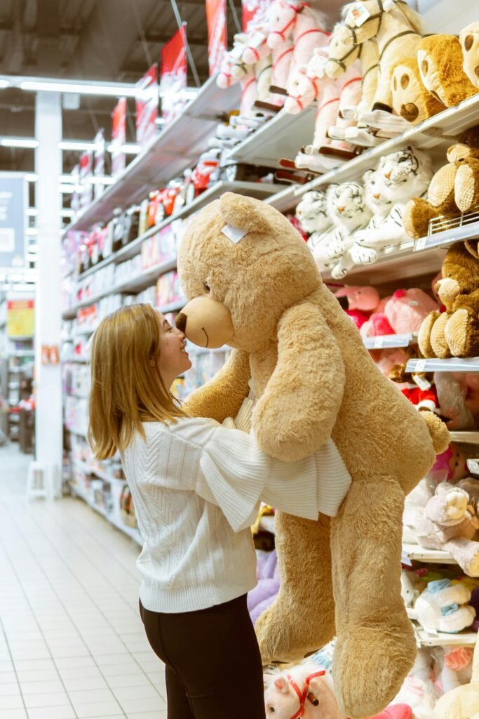 A woman chooses a large teddy bear in a retail toy section, surrounded by stuffed animals.