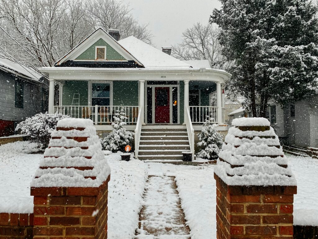 A picturesque bungalow with a snow-covered roof and trees depicting a serene winter scene.