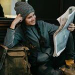 Casually dressed woman enjoying reading a newspaper while sitting with a backpack indoors, exuding a relaxed vibe.