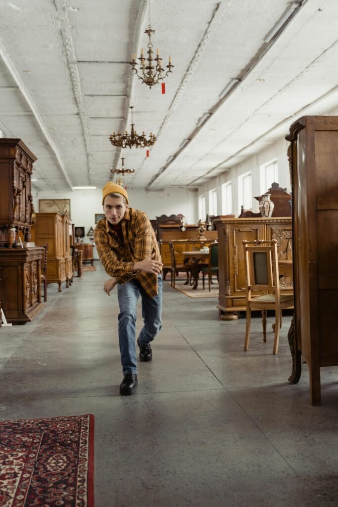 A young man casually poses in a vintage furniture shop, surrounded by antique wooden pieces.