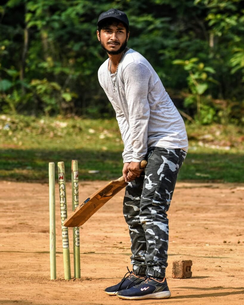 Man holding a cricket bat in batting position on a rustic field with stumps.
